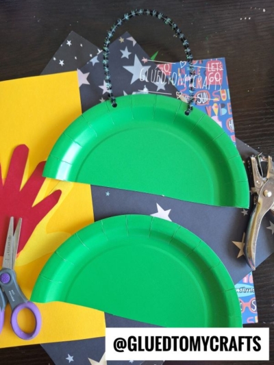 Two green paper plates cut in half with holes punched at the top, one connected by a black pipe cleaner handle. Scissors, a red cut-out hand, yellow and black paper, and a hole punch are nearby on a dark surface.