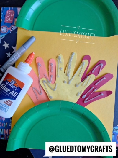 A craft project featuring handprint cutouts glued to yellow paper to form a turkey, with green paper plates as wings, a bottle of Elmers glue, a silver marker, and the @gluedtomycrafts handle displayed.