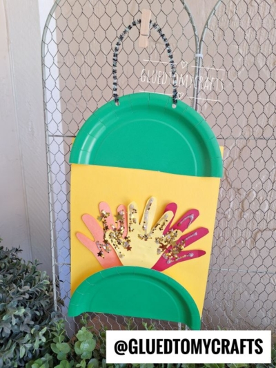 A colorful summer craft featuring cut-out paper hands in fall colors on yellow paper, with green paper plate halves as a border, hanging on a wire fence. Text reads @GLUEDTOMYCRAFTS.