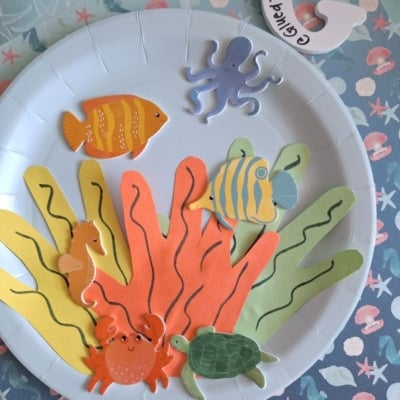 A paper plate decorated as an underwater scene with colorful paper seaweed, fish, a seahorse, a crab, a turtle, and an octopus cutout on the side. @GLUEDTOMYCRAFTS is labeled at the bottom.