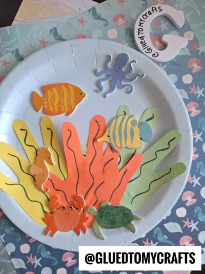 A paper plate decorated as an underwater scene with colorful paper seaweed, fish, a seahorse, a crab, a turtle, and an octopus cutout on the side. @GLUEDTOMYCRAFTS is labeled at the bottom.