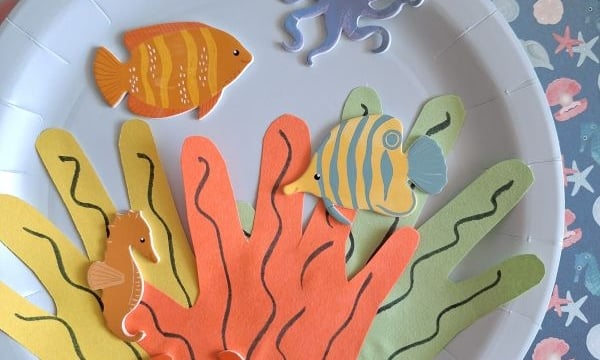 A paper plate decorated as an underwater scene with colorful paper seaweed, fish, a seahorse, a crab, a turtle, and an octopus cutout on the side. @GLUEDTOMYCRAFTS is labeled at the bottom.