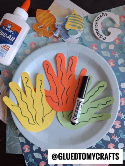 A paper plate holds yellow, orange, and green cutouts shaped like hands with squiggly lines, a black marker, Elmer’s glue, ocean animal cutouts, and a circular @GLUEDTOMYCRAFTS logo on a craft table.
