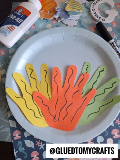 A paper plate holds cutouts of hand shapes in orange, yellow, and green with squiggly lines, resembling seaweed or coral. Glue and markers are nearby. The text @GLUEDTOMYCRAFTS appears at the bottom.