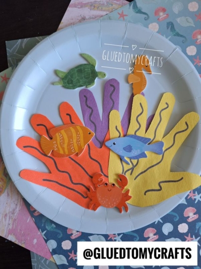 A paper plate aquarium decorated with colorful paper cutouts of sea creatures, coral, and hand shapes. Fish, a turtle, a crab, and coral are featured on the plate, with the handle @GLUEDTOMYCRAFTS at the bottom.