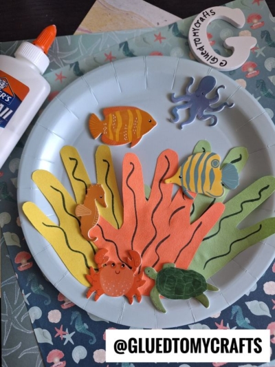 A paper plate decorated with colorful paper seaweed and cut-out sea animals, including a crab, turtle, octopus, seahorse, and fish. Elmer’s glue and craft materials are visible nearby. @GLUEDTOMYCRAFTS is labeled at the bottom.