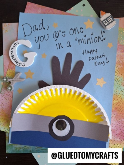 A handmade HANDPRINT FATHER'S DAY card features a minion design with a yellow painted half paper plate, black and gray paper details, and a sweet message: Dad, you are one in a minion! Happy Fathers Day! Craft supplies are scattered around.