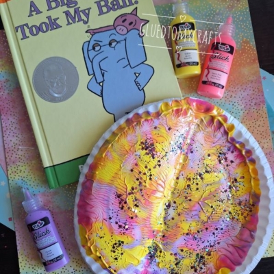 Children’s book “A Big Guy Took My Ball!” by Mo Willems, next to a painted paper plate craft in pink, yellow, and white, with black dots, and three bottles of puffy paint on a colorful surface.