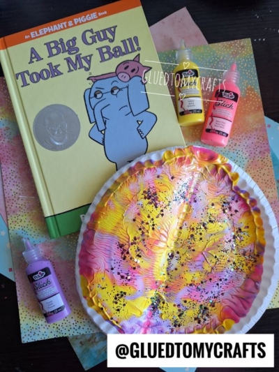 Children’s book “A Big Guy Took My Ball!” by Mo Willems, next to a painted paper plate craft in pink, yellow, and white, with black dots, and three bottles of puffy paint on a colorful surface.