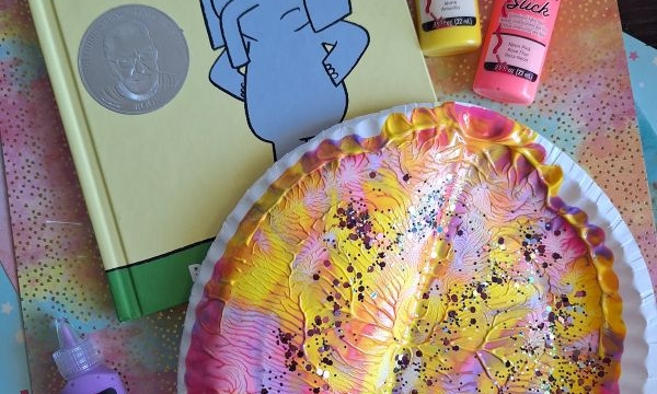 Children’s book “A Big Guy Took My Ball!” by Mo Willems, next to a painted paper plate craft in pink, yellow, and white, with black dots, and three bottles of puffy paint on a colorful surface.