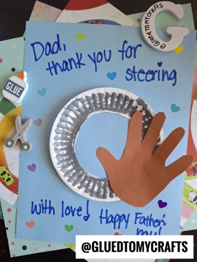 A handmade Handprint Father's Day card uses a paper plate as a steering wheel and a brown paper handprint "holding" it. The card reads, Dad, thank you for steering with love! Happy Father's Day! Craft supplies are nearby.