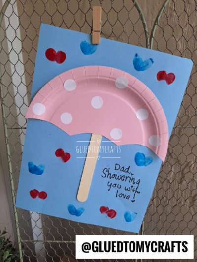 A handmade HANDPRINT FATHER'S DAY card features a pink paper plate umbrella with white dots, a wooden stick handle, and red and blue hearts on blue paper. Text reads, Dad, Showering you with love! The handle includes @GLUEDTOMYCRAFTS.