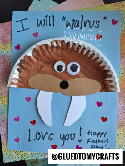 A handmade Father’s Day card features a walrus made from a painted paper plate, googly eyes, and tusks on blue paper—perfect for a HANDPRINT FATHER'S DAY craft—with “I will walrus love you! Happy Father’s Day!” written on it.