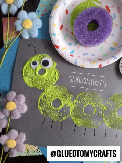 A Pool Noodle Stamped Caterpillar craft made with green paint circles, googly eyes, and drawn antennae on black paper. A purple sponge, paper plate, and felt flowers are nearby. The text @GLUETOMYCRAFTS is on the image.