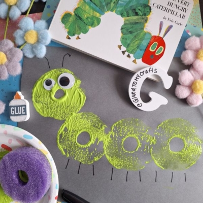 A Pool Noodle Stamped Caterpillar craft made with green and yellow paint circles, googly eyes, and a purple sponge on black paper. The Very Hungry Caterpillar book, pastel pom-poms, glue, and a clay “G” are nearby. @GLUEDTOMYCRAFTS is labeled.