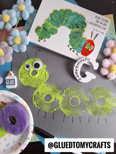A Pool Noodle Stamped Caterpillar craft made with green and yellow paint circles, googly eyes, and a purple sponge on black paper. The Very Hungry Caterpillar book, pastel pom-poms, glue, and a clay “G” are nearby. @GLUEDTOMYCRAFTS is labeled.