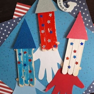 Three popsicle stick rockets with glittery paper tops and star stickers are arranged on handprint cutouts over blue and American flag-themed paper. An Instagram handle, @GLUEDTOMYCRAFTS, is shown at the bottom.