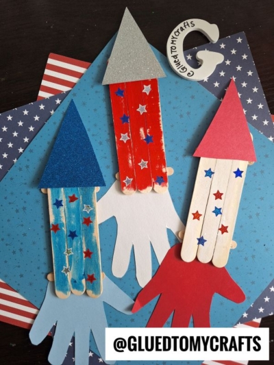 Three popsicle stick rockets with glittery paper tops and star stickers are arranged on handprint cutouts over blue and American flag-themed paper. An Instagram handle, @GLUEDTOMYCRAFTS, is shown at the bottom.