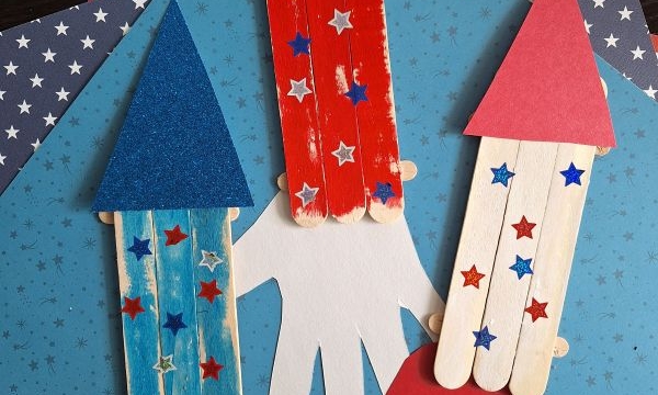 Three popsicle stick rockets with glittery paper tops and star stickers are arranged on handprint cutouts over blue and American flag-themed paper. An Instagram handle, @GLUEDTOMYCRAFTS, is shown at the bottom.