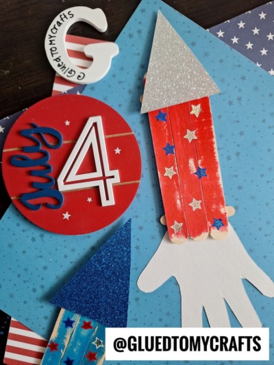 Handmade July 4th crafts, including a red July 4 circle and a popsicle stick rocket decorated with stars and glitter, on blue paper with the Instagram handle @GLUEDTOMYCRAFTS.