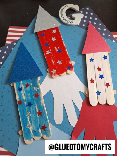 Three rocket crafts made from colored popsicle sticks and paper triangles are displayed on patriotic backgrounds with red, white, and blue stars. A @gluedtomycrafts tag appears in the bottom corner.