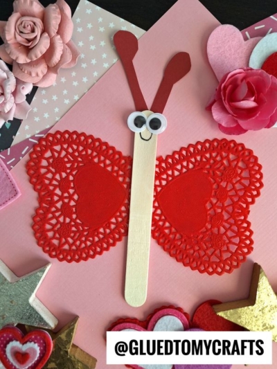 A butterfly craft made from red paper doilies, a wooden popsicle stick, googly eyes, and heart-shaped antennae, surrounded by pink and red flowers and decorative shapes.