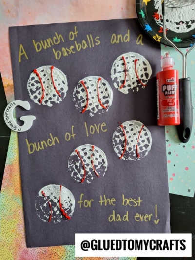 A potato masher stamped craft on black paper with white and red painted baseballs, handwritten text saying A bunch of baseballs and a bunch of love for the best dad ever! Various art supplies are scattered around.