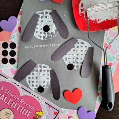 Valentine’s Day dog craft on black paper using white paint, black paper ears, heart cutouts, and a paint roller. A Charlie Brown Valentine book is in the corner. ©GLUEDTOMYCRAFTS is written on the paper.