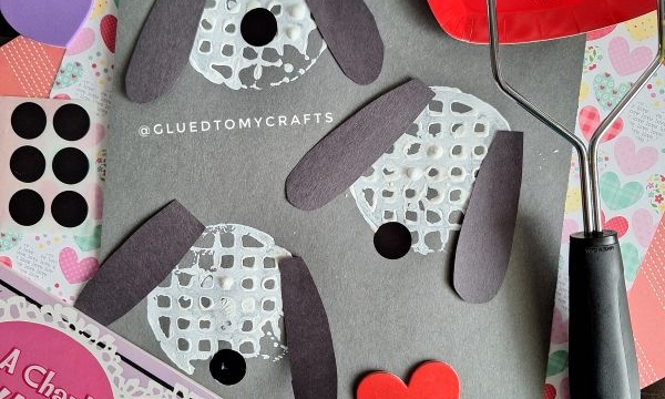Valentine’s Day dog craft on black paper using white paint, black paper ears, heart cutouts, and a paint roller. A Charlie Brown Valentine book is in the corner. ©GLUEDTOMYCRAFTS is written on the paper.