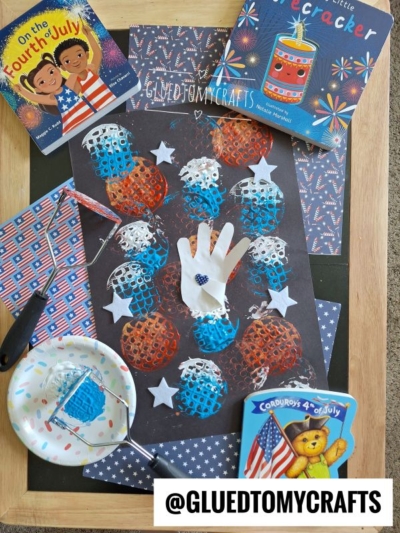 A Fourth of July-themed flat lay with patriotic children’s books, a star-patterned napkin, a pan with confetti, and a potato masher stamped craft featuring red, white, and blue paint, stars, and a handprint on black paper.