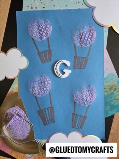A blue paper features three hot air balloon shapes created with purple potato masher stamped craft techniques; decorative clouds border the edges, and @GLUEDTOMYCRAFTS is displayed at the bottom.