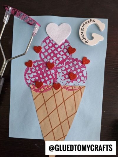 A potato masher ice cream cone craft featuring a cone with three scoops, decorated with red glitter hearts and a white heart. The pink-handled masher and “G” label @GLUEDTOMYCRAFTS sit nearby on a blue background.