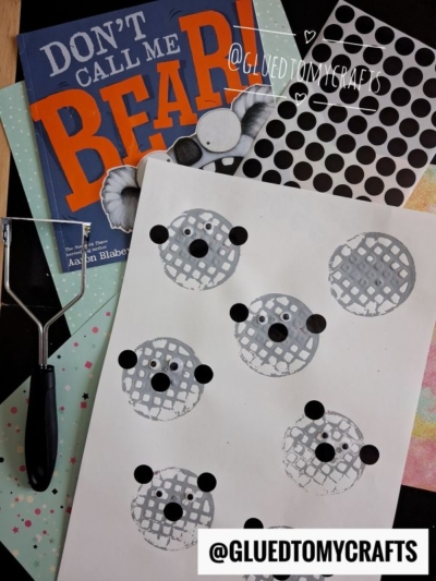 A children’s craft setup featuring the book “Don’t Call Me Bear!”, a brayer, black sticker dots, and sheet with potato masher stamped bear faces, created from gray circles, black dots, and lattice patterns. @GLUEDTOMYCRAFTS watermark is present.