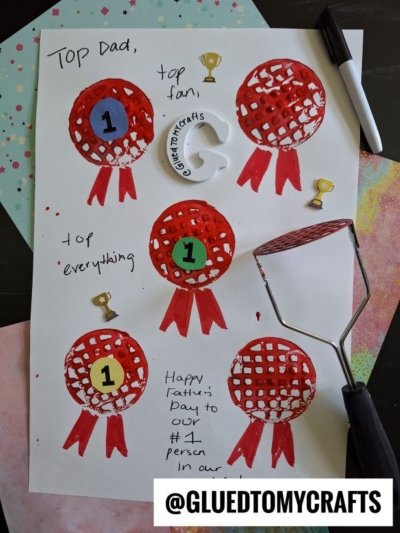 A handmade Fathers Day card features five red, ribbon-like 1st place awards, gold trophies, handwritten notes like Top Dad, and a creative potato masher stamped craft. A paint roller and palette are nearby. @GLUEDTOMYCRAFTS.