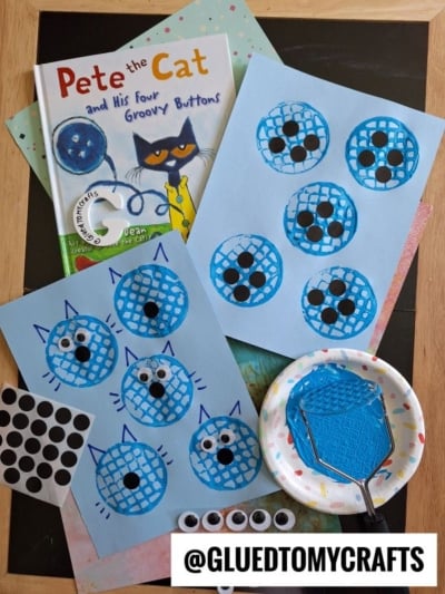 Two blue papers feature cat faces made with a potato masher stamped craft, blue paint, and black buttons, inspired by Pete the Cat and His Four Groovy Buttons. Supplies also include googly eyes, stickers, and a small plate with blue paint.