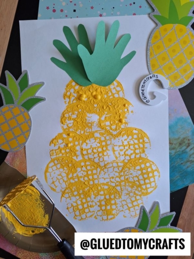 A pineapple craft made with yellow paint in an oval shape using a potato masher stamped craft technique, topped with green paper cutout leaves and surrounded by pineapple decorations. The handle of a potato masher and a “@GLUEDTOMYCRAFTS” label are visible.