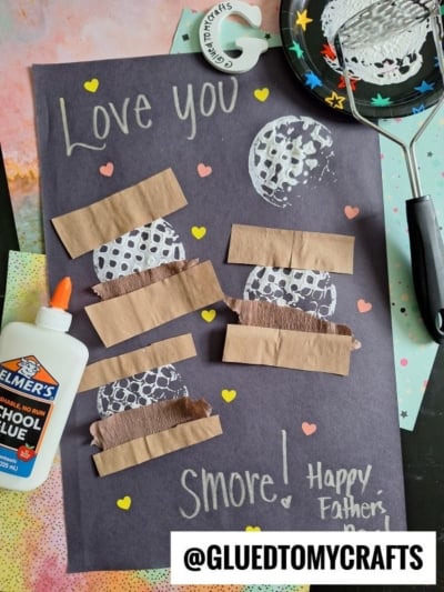 A handmade Fathers Day card on black paper with Love you Smore! in yellow, decorated with potato masher stamped smores, hearts, white glue, a spatula, and a lunar-themed background. @GLUEDTOMYCRAFTS is at the bottom.