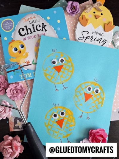 A blue paper features three potato masher stamped chicks made from textured paint and googly eyes, surrounded by artificial flowers, a spring sign with a chick, the children’s book "There's a Little Chick in Your Book," and craft supplies.