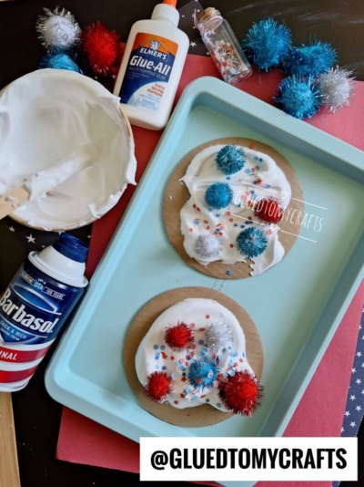 Two round cardboard pieces on a blue tray are decorated with white foam, red, blue, and silver pom-poms, and star-shaped confetti—perfect for puffy paint crafts. Shaving cream, glue, and glitter glue are shown. Text reads @GLUEDTOMYCRAFTS.
