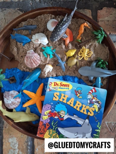 A wooden tray filled with sand, seashells, plastic sea animals, and a Dr. Seuss Sharks book—perfect for story time sensory bin ideas; @GLUEDTOMYCRAFTS is printed at the bottom.