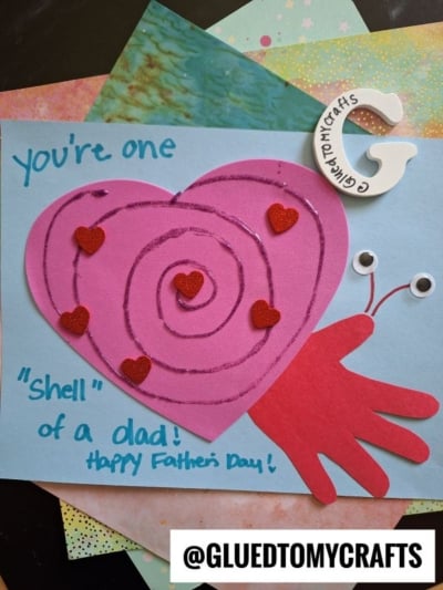 Handmade HANDPRINT FATHER'S DAY card featuring a snail made from a pink heart shell, red glitter hearts, and a red handprint body. Text reads: You’re one shell of a dad! Happy Father’s Day!.
