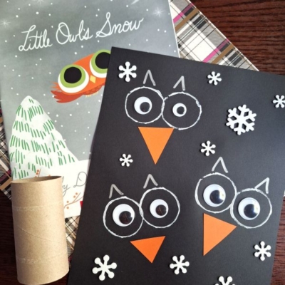 A children’s owl craft made with a black paper background, white and orange paper pieces, and snowflake cutouts sits next to the book “Little Owl’s Snow” and a cardboard tube.