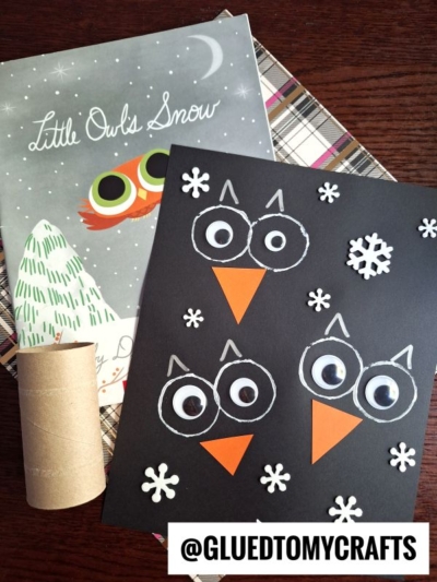 A children’s owl craft made with a black paper background, white and orange paper pieces, and snowflake cutouts sits next to the book “Little Owl’s Snow” and a cardboard tube.