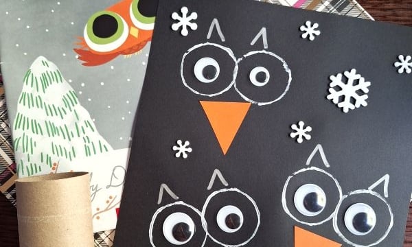 A children’s owl craft made with a black paper background, white and orange paper pieces, and snowflake cutouts sits next to the book “Little Owl’s Snow” and a cardboard tube.