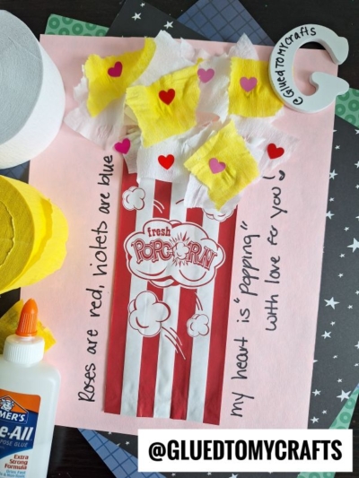 A handmade popcorn-themed Valentine’s card features a popcorn bag cutout with crepe paper popcorn craft, heart stickers, and the message: Roses are red, violets are blue, my heart is popping with love for you.