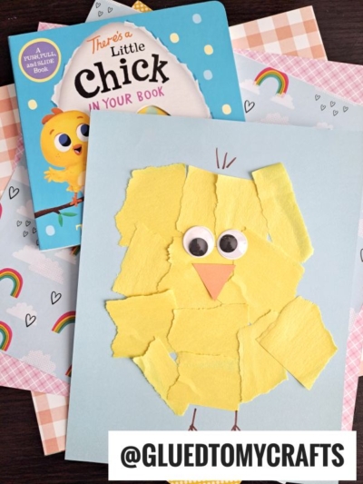 A children’s craft project features a torn paper baby chick with googly eyes on blue paper, lying on pastel rainbow-patterned sheets near a chick-themed children’s book.