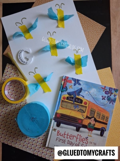 A butterfly craft project with tissue paper, tape, pipe cleaners, and a book titled Butterflies on the First Day of School displayed on patterned paper sheets—perfect for sparking creative INSECT CRAFT IDEAS in young learners.