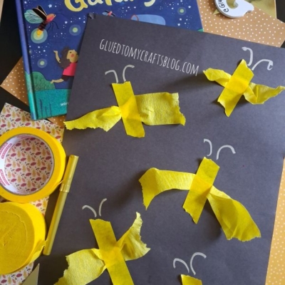 A childrens book called Firefly Galaxy sits near a black paper with yellow tissue paper fireflies. Craft supplies like tape and scissors are scattered around. @Glutetomycrafts is printed on the photo.