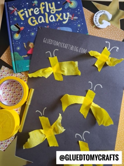 A childrens book called Firefly Galaxy sits near a black paper with yellow tissue paper fireflies. Craft supplies like tape and scissors are scattered around. @Glutetomycrafts is printed on the photo.