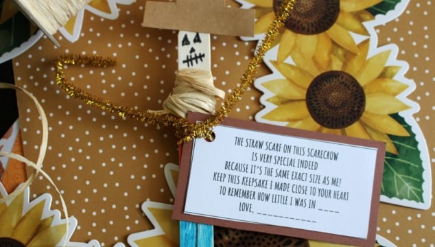 A handmade scarecrow craft with a note attached, decorated with sunflower patterns. The note explains the significance of the scarecrows scarf and has a space for a childs name.